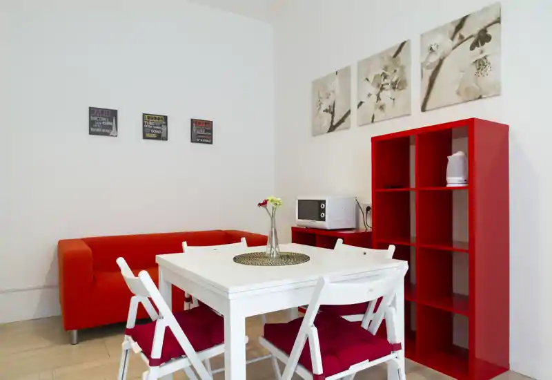 Coin salon/salle à manger lumineux et moderne avec table blanche, canapé et étagères rouges, créant un espace accueillant pour petits groupes.