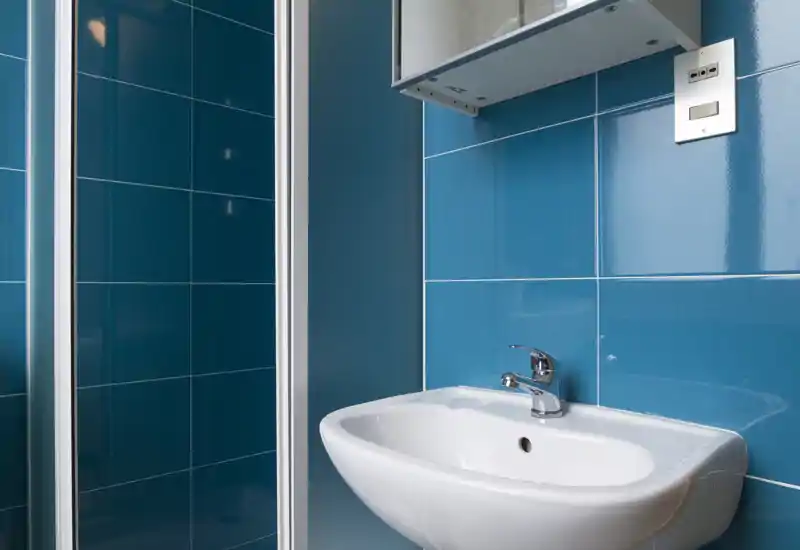 Salle de bains moderne et compacte avec carreaux bleus brillants, lavabo mural blanc et robinet chromé à côté d'une cabine de douche encadrée.