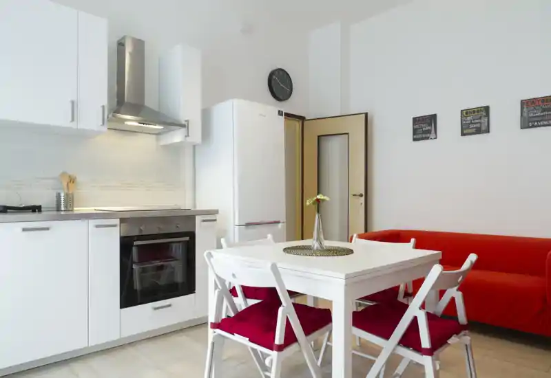 Kitchenette lumineuse avec placards blancs, four moderne et une petite table à manger avec chaises à coussins rouges à côté d'un canapé rouge — espace de vie et de cuisson compact et élégant.