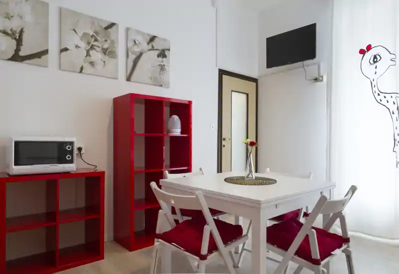 Coin repas moderne et lumineux avec une table blanche et quatre chaises, rehaussé par des coussins rouges et des étagères assorties; la lumière naturelle filtrée par les voilages apporte une ambiance accueillante.