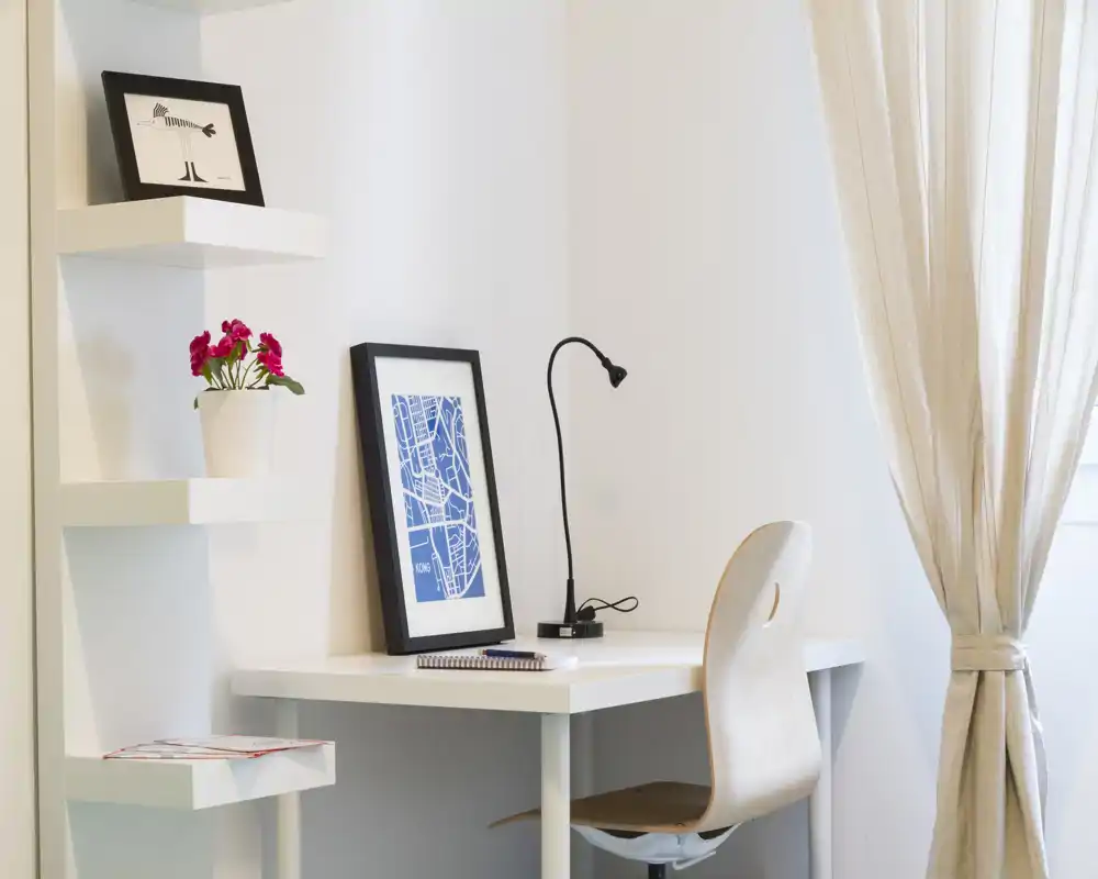 Espacio de trabajo luminoso y minimalista con escritorio blanco, silla de madera, lámpara de mesa y cuadros junto a una ventana con cortina clara.