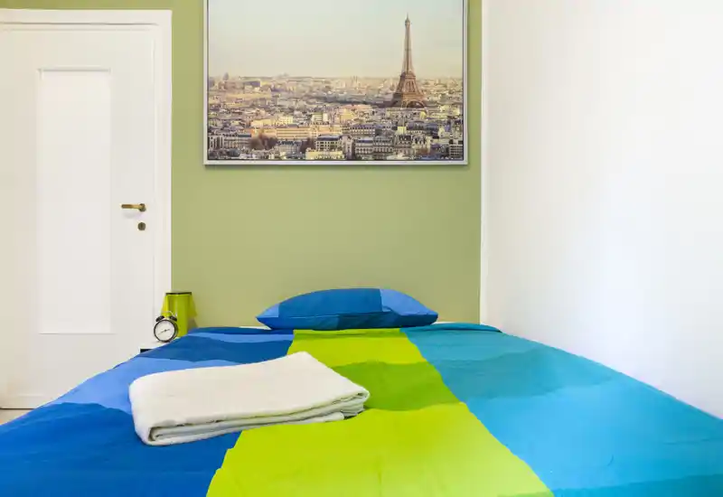 Dormitorio compacto y colorido con cama individual bien hecha, ropa de cama azul y verde, almohada y toallas dobladas; cuadro de la ciudad sobre la cama y luz suave y agradable.