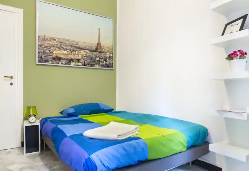 Dormitorio compacto y luminoso con cama doble con ropa de cama azul y verde, mesita con lámpara y despertador, cuadro en la pared y estantes con planta — ideal para una estancia corta y acogedora.