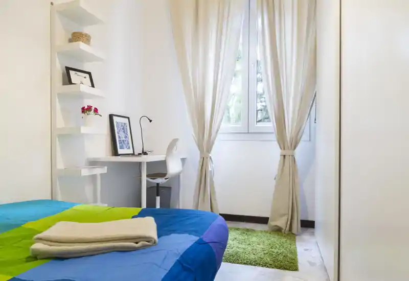 Dormitorio luminoso y acogedor con cama de colores, mantas dobladas y un escritorio junto a la ventana que deja entrar luz natural.