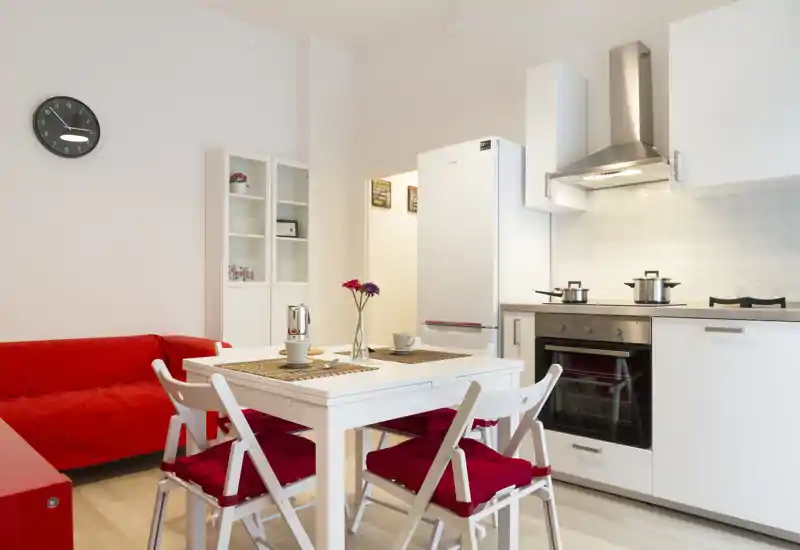 Cocina moderna y luminosa con mesa blanca, sillas con cojines rojos y frigorífico y horno compactos — ideal para un apartamento urbano acogedor.