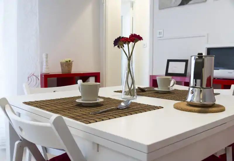 Área de comedor luminosa y moderna con mesa blanca para dos, manteles individuales, una cafetera y un jarrón con flores: estilo limpio y minimalista perfecto para anuncios de pisos pequeños.