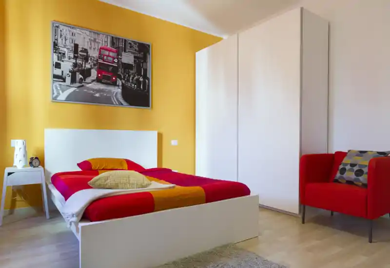 Camera da letto moderna e luminosa con letto bianco a contenitore, biancheria rossa e arancione e una poltrona accogliente. Un grande armadio e un quadro completano l'ambiente piacevole e ben illuminato.