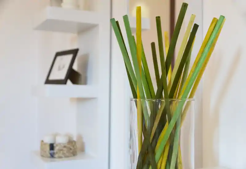 Dettaglio luminoso di un soggiorno moderno con un vaso di vetro decorativo contenente steli verdi e gialli; mensole bianche e piccoli oggetti (candele, cornice) sullo sfondo.