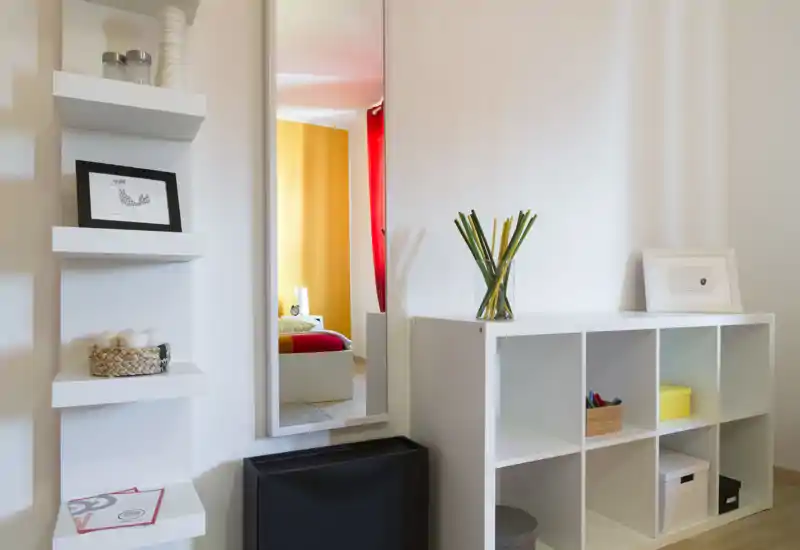 Angolo di camera luminoso e moderno con una libreria bianca e uno specchio alto che riflette un letto colorato e una parete accentuata gialla.