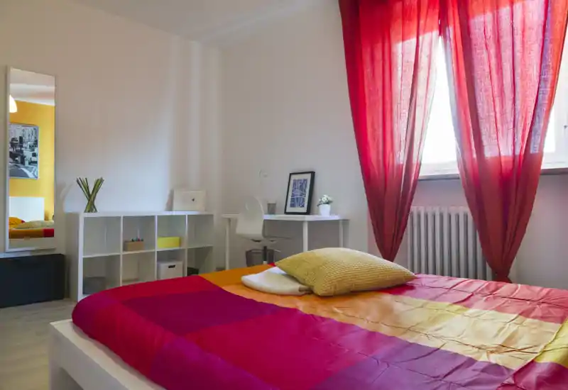 Camera da letto luminosa e colorata con letto ampio, tessuti caldi e una piccola scrivania vicino alla finestra — perfetta per riposare e lavorare.