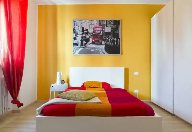 Camera da letto luminosa e colorata con letto matrimoniale al centro su parete gialla, biancheria rosso-arancione, comodino e armadio — spazio accogliente e ben illuminato ideale per annunci.