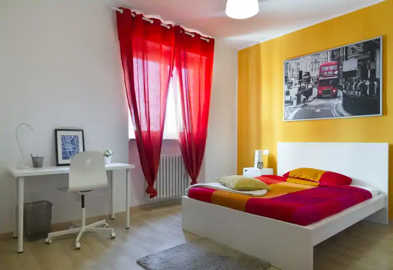 Camera da letto luminosa e colorata con letto bianco, biancheria rosso-arancio e parete accentata gialla; una scrivania vicino alla finestra la rende adatta anche per studio o lavoro.