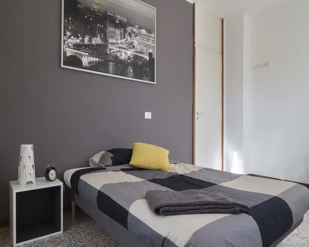 Camera da letto ben arredata con piumone grigio a motivo, cuscino giallo come elemento d'accento e comodino con lampada e sveglia; arredi neutri e luce naturale soffusa.