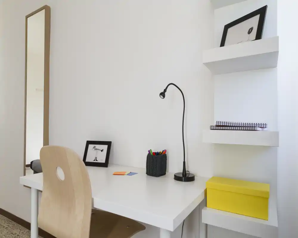 Angolo studio luminoso e minimale con scrivania bianca, sedia in legno, lampada da tavolo e accessori ordinati — un compatto spazio lavoro domestico.