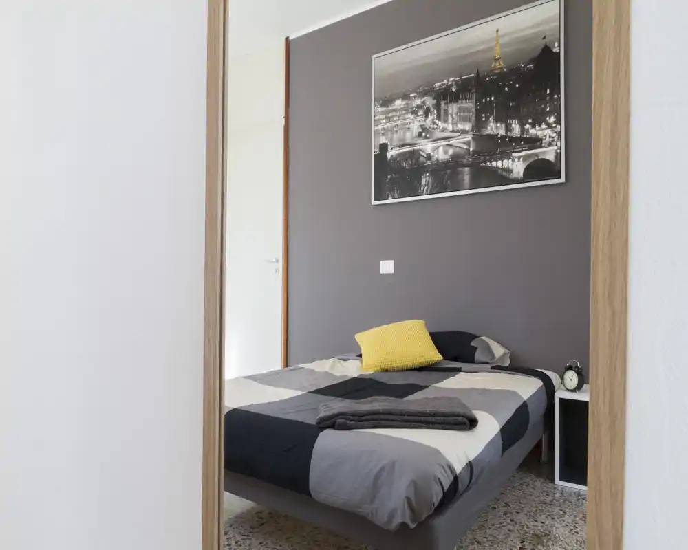 Camera luminosa e ordinata con letto matrimoniale, cuscino giallo decorativo e quadro sulla parete. Buona luce naturale e arredo minimale, perfetta per annunci di affitto.
