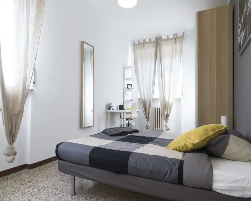 Camera da letto luminosa e moderna con letto matrimoniale coperto da una trapunta grigia a quadri, cuscino giallo come tocco di colore, armadio alto e una piccola scrivania vicino alla finestra — ariosa e semplicemente arredata.