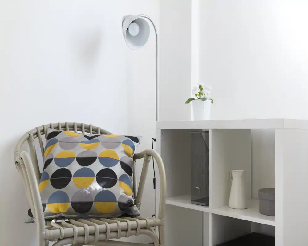 Angolo soggiorno luminoso e minimal con una poltrona intrecciata, cuscino colorato a fantasia, lampada da terra e una libreria bianca con oggetti decorativi.