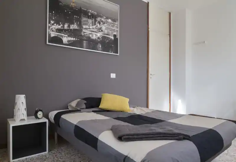 Camera da letto ben arredata con piumone grigio a motivo, cuscino giallo come elemento d'accento e comodino con lampada e sveglia; arredi neutri e luce naturale soffusa.