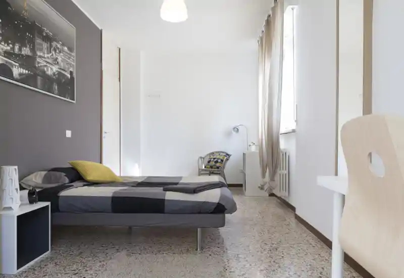 Camera da letto luminosa e moderna con letto matrimoniale con biancheria geometrica, comodino, sedia e luce naturale da una grande finestra — ideale per annunci che puntano su design pulito e comfort.