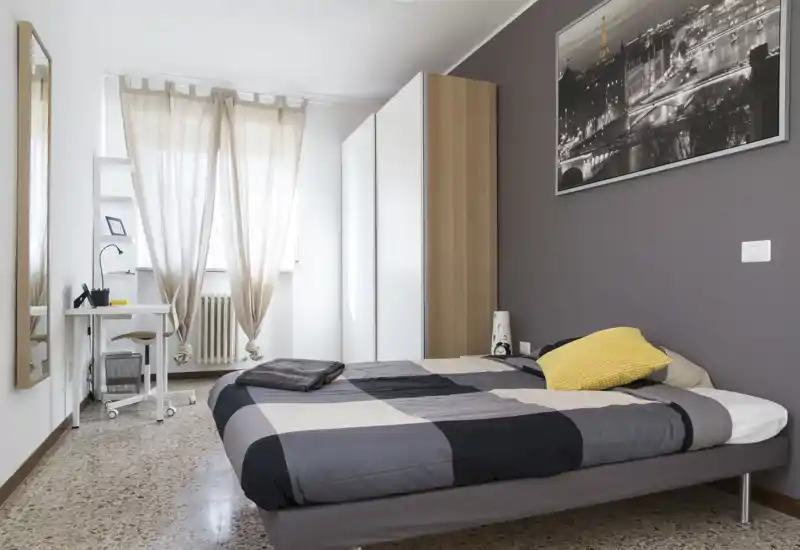 Camera da letto luminosa e ben arredata con letto matrimoniale, armadio e scrivania vicino alla finestra. La luce naturale e l’arredo sobrio creano un’atmosfera accogliente, perfetta per annunci.