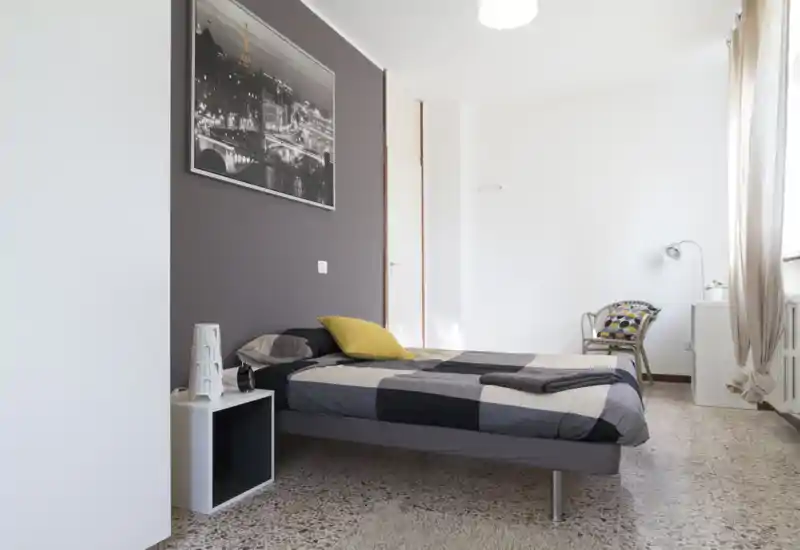 Camera da letto luminosa e moderna con letto matrimoniale con copriletto grigio a blocchi, comodino con lampada e finestra che illumina la stanza con luce naturale.