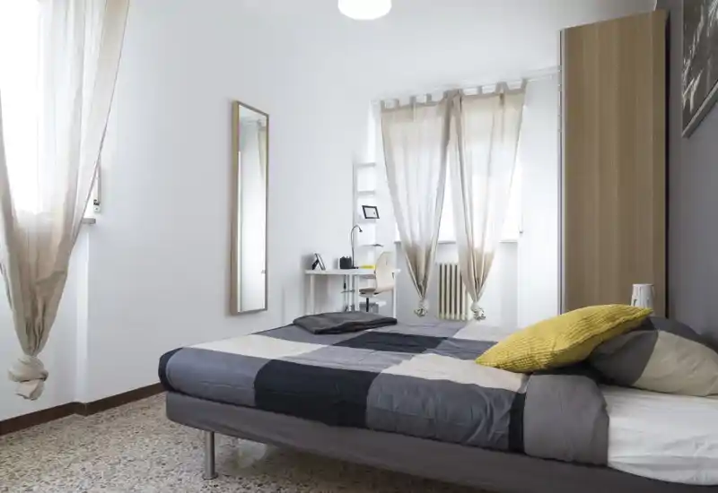 Camera da letto luminosa e moderna con letto matrimoniale coperto da una trapunta grigia a quadri, cuscino giallo come tocco di colore, armadio alto e una piccola scrivania vicino alla finestra — ariosa e semplicemente arredata.