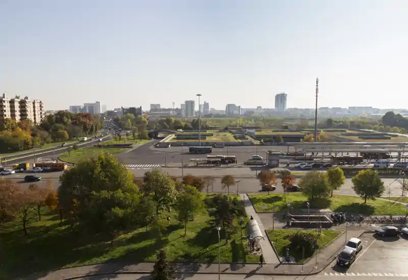 Vue extérieure ensoleillée sur un parc et les routes adjacentes avec des bus et la silhouette de la ville au loin — idéale pour mettre en valeur le quartier et les liaisons.