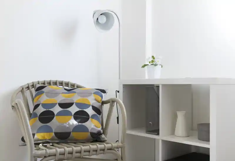 Coin salon lumineux et minimaliste avec un fauteuil en osier, un coussin à motifs colorés, une lampe sur pied et une étagère blanche avec objets décoratifs.