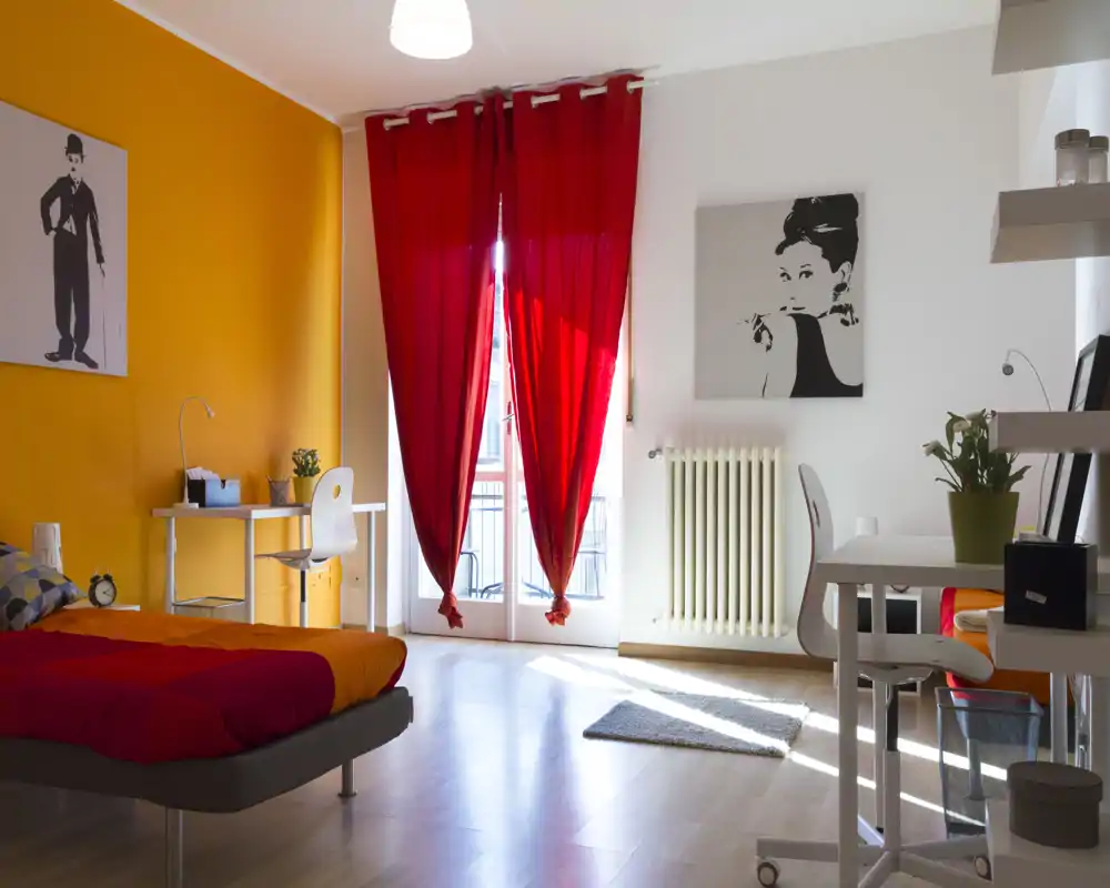 Camera da letto luminosa e colorata con letto matrimoniale, scrivania e grande finestra con tende rosse che lasciano entrare luce naturale; stile moderno e accogliente ideale per annunci.