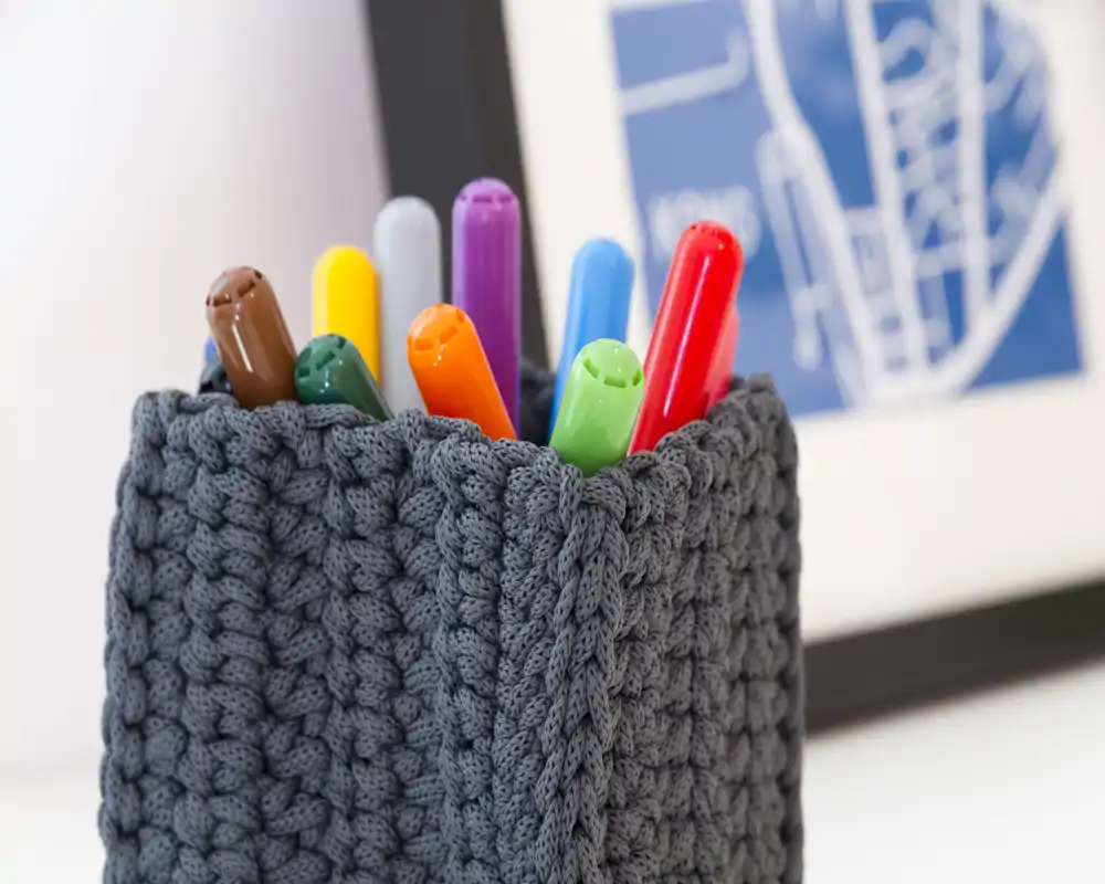 Primo piano di un porta‑penne lavorato a maglia con pennarelli colorati su una superficie pulita; scatto dettagliato incentrato sulla trama e sulla cancelleria.