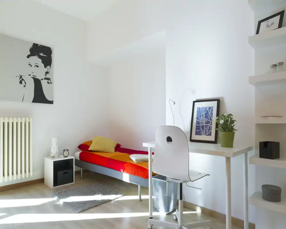 Camera singola moderna e luminosa con letto rosso e arancione, scrivania compatta e mensole — ideale per uno studente o un inquilino singolo. Abbondante luce naturale e arredi minimal rendono l’ambiente accogliente.