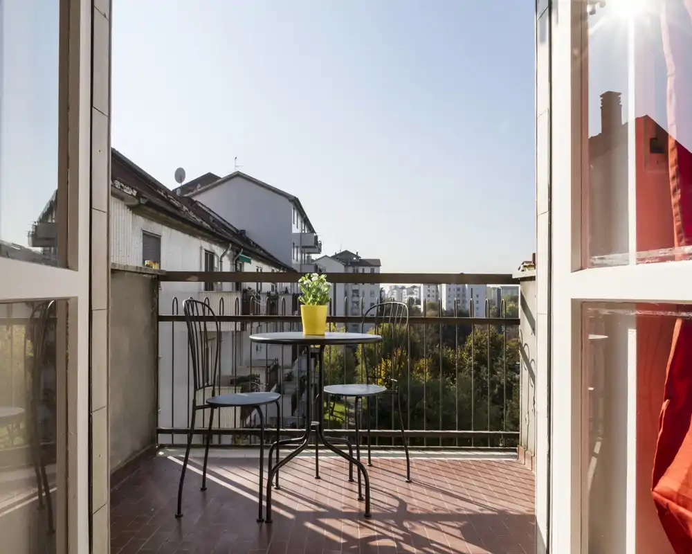 Balcone soleggiato con un piccolo tavolo rotondo, tre sedie in metallo e una pianta in vaso, con vista sugli edifici vicini e sul verde.