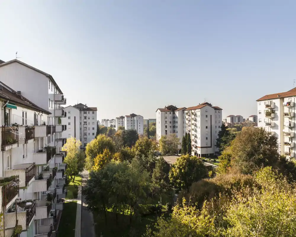 Vista soleggiata da un balcone su un complesso residenziale con cortili verdi e palazzine di media altezza sotto un cielo sereno.