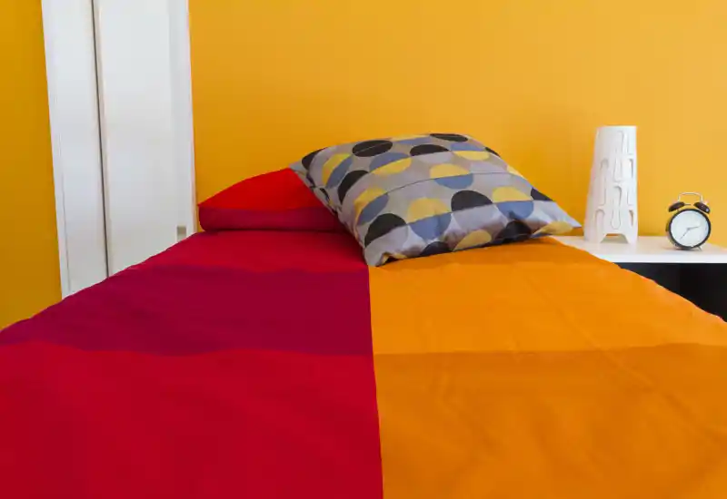 Camera da letto singola, luminosa e moderna con biancheria colorata e cuscino decorativo su un letto ordinato davanti a una parete gialla; comodino con lampada e sveglia.
