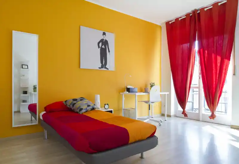 Camera da letto moderna e luminosa con letto matrimoniale in biancheria rossa e arancione, una scrivania semplice vicino alla finestra e una parete gialla a contrasto che illumina l’ambiente.