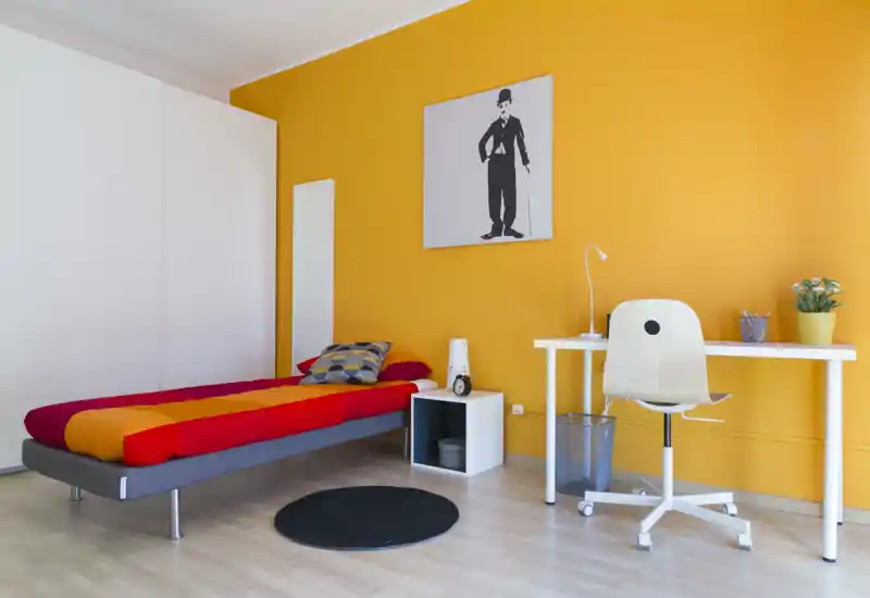 Camera singola moderna e luminosa con letto basso e copriletto colorato, scrivania ordinata e parete accentata arancione — perfetta per uno studente o un single.