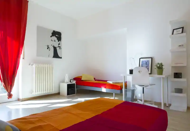 Camera da letto luminosa e colorata con due letti visibili (uno in primo piano e un singolo sul fondo), biancheria rossa e arancione, piccola scrivania e scaffale – ideale per un soggiorno comodo per due o famiglia.