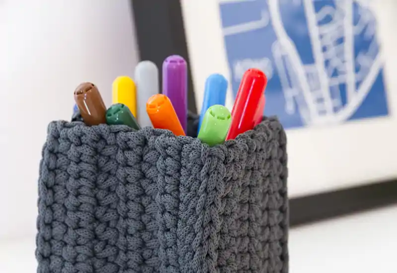 Primo piano di un porta‑penne lavorato a maglia con pennarelli colorati su una superficie pulita; scatto dettagliato incentrato sulla trama e sulla cancelleria.