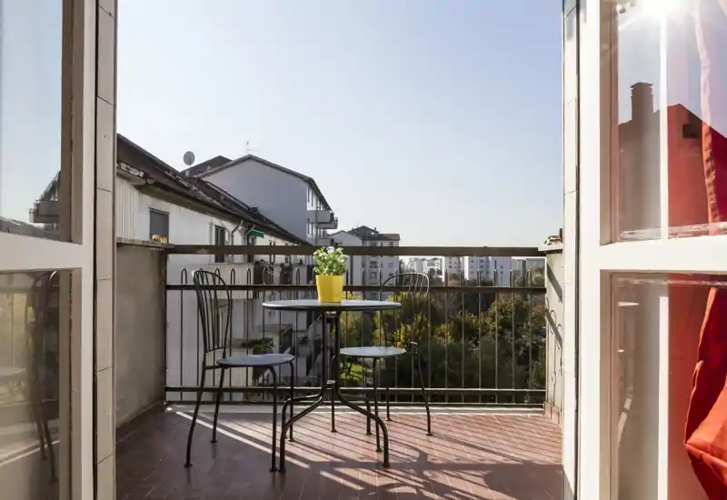 Balcone soleggiato con un piccolo tavolo rotondo, tre sedie in metallo e una pianta in vaso, con vista sugli edifici vicini e sul verde.