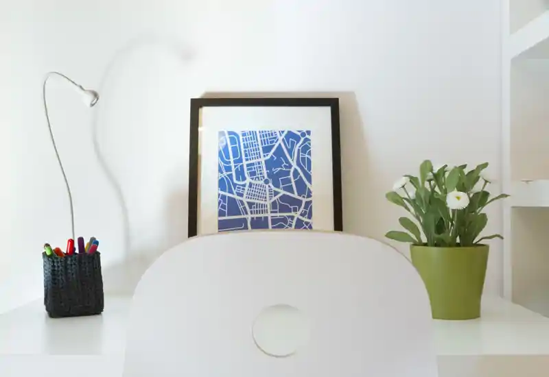 Angolo scrivania luminoso e ordinato con sedia bianca, stampe incorniciate blu e una pianta in vaso — un tranquillo spazio di lavoro in soggiorno o monolocale.