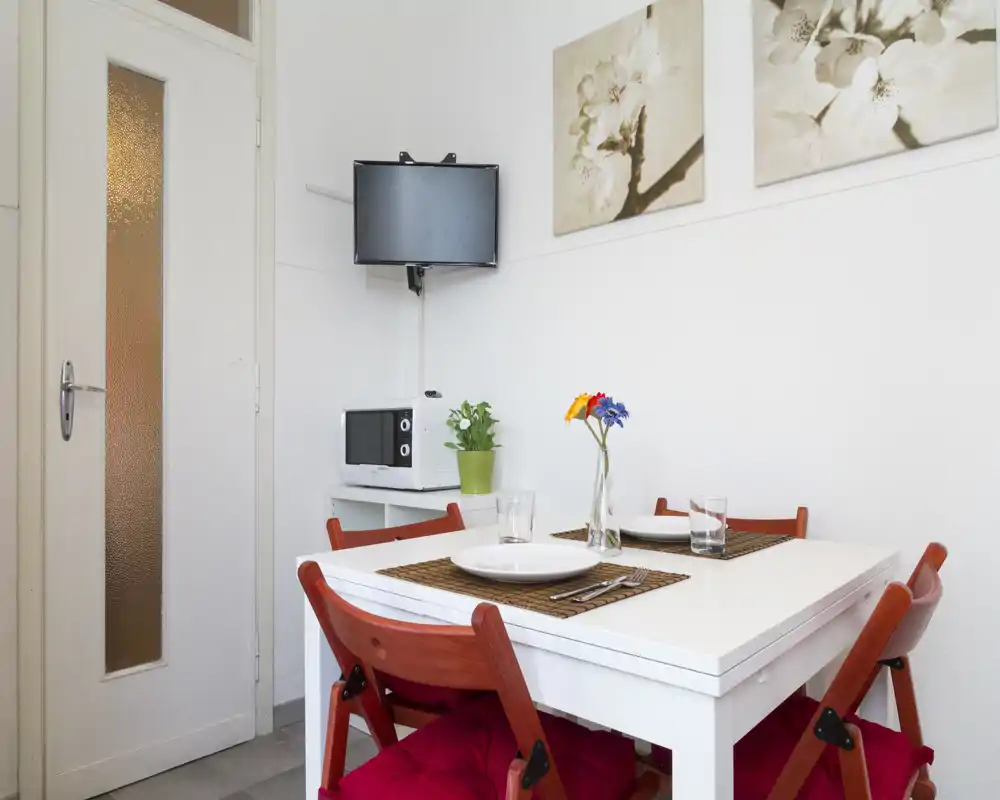 Zona pranzo luminosa e ben arredata con tavolo bianco apparecchiato, sedie con cuscini rossi, tovagliette decorative e un piccolo vaso di fiori. Si vedono elementi di angolo cottura (microonde, TV) e pareti bianche pulite.