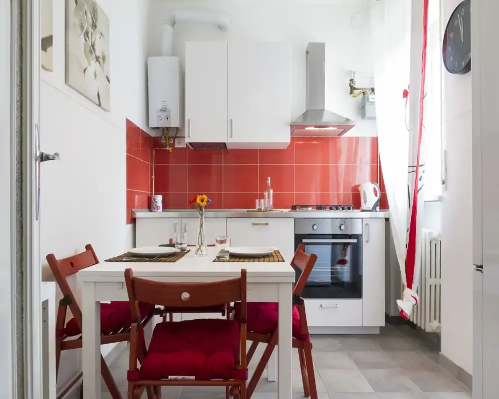 Cucina compatta e luminosa con un piccolo tavolo da pranzo per due. Mobiletti bianchi, paraschizzi rosso e elettrodomestici in acciaio inox per un aspetto moderno e accogliente.
