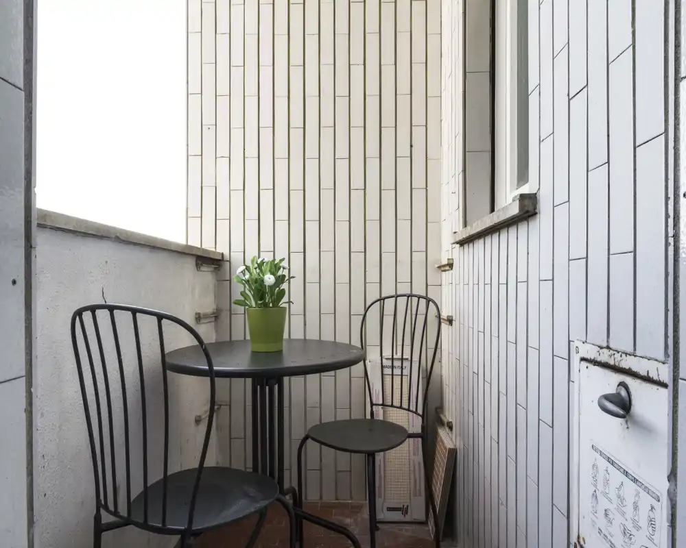 Piccolo balcone con tavolino rotondo in metallo, due sedie abbinate e una pianta in vaso; pareti piastrellate e spazio compatto perfetto per un caffè mattutino.