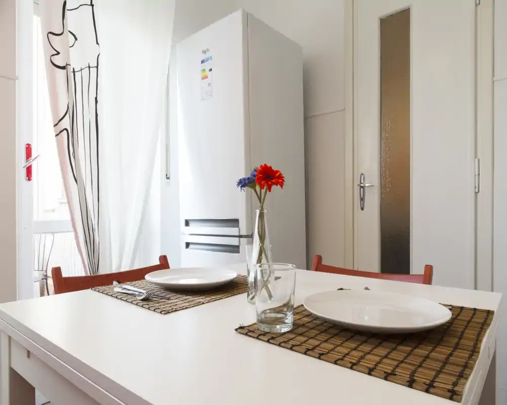 Angolo cucina luminoso e compatto con un tavolo bianco apparecchiato per due, frigorifero visibile e un piccolo vaso di fiori che crea un’atmosfera accogliente.