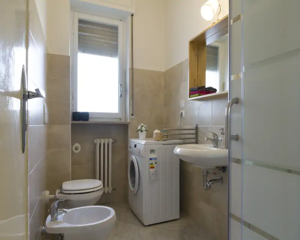 Bagno luminoso e ordinato con wc, bidet, lavabo a parete, doccia con porta in vetro satinato e lavatrice sotto la finestra.
