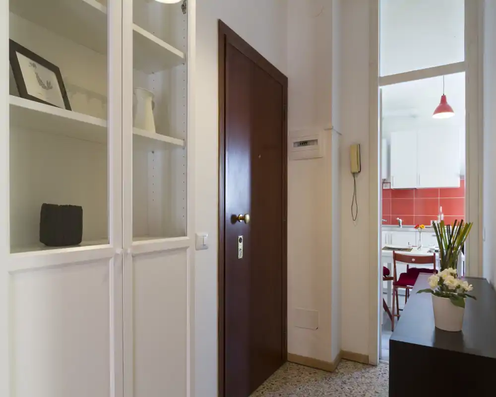 Ingresso ordinato e ben illuminato con porta d'ingresso in legno massiccio, armadi bianchi incorporati e vista sulla cucina, ideale per una buona prima impressione.