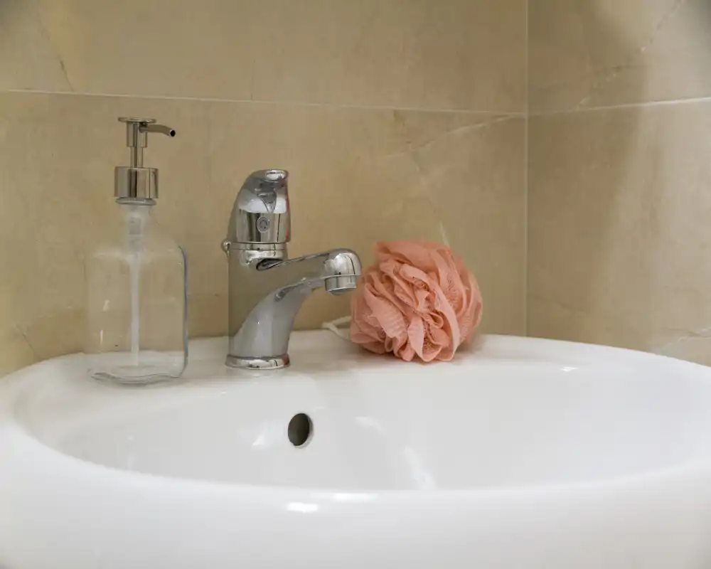 Primo piano di un lavabo bagno pulito con rubinetto cromato, dispenser di sapone liquido e spugna rosa su piastrelle.