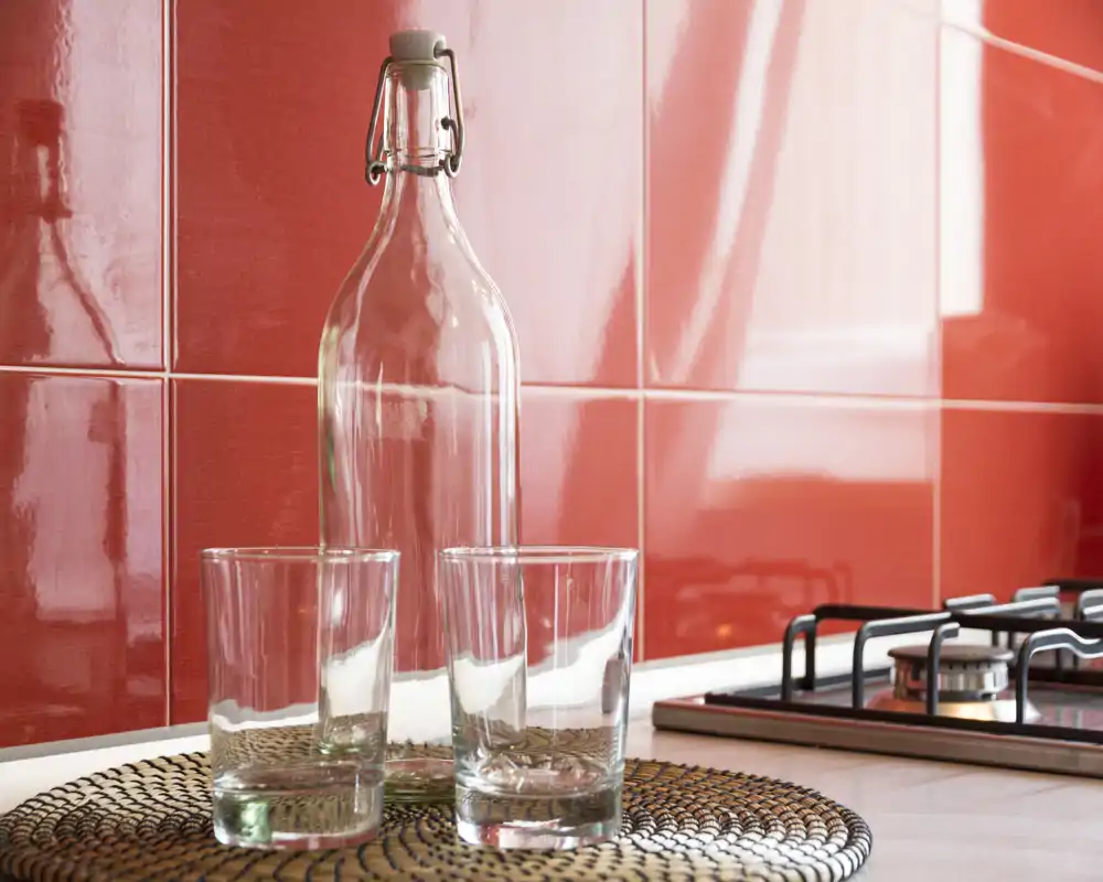 Bancone cucina luminoso e moderno con bicchieri decorativi e una bottiglia trasparente davanti a piastrelle rosse lucide e un piano cottura a gas — uno scatto dettagliato che mette in risalto piano e rivestimento.