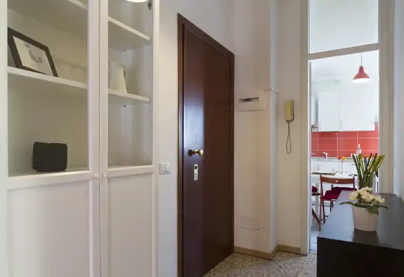 Ingresso ordinato e ben illuminato con porta d'ingresso in legno massiccio, armadi bianchi incorporati e vista sulla cucina, ideale per una buona prima impressione.