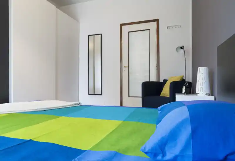 Bright, modern bedroom with a colorful bedspread occupying the foreground, a wardrobe and a tall mirror along the wall, plus a bedside table and armchair.